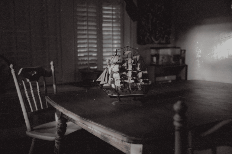 A dimly lit room with a wooden table and chairs, where a detailed model ship sits bathed in soft, slatted light—evoking memory, loss, and the quiet presence of what’s gone.