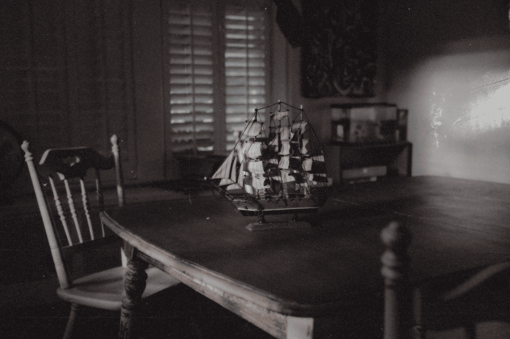 A dimly lit room with a wooden table and chairs, where a detailed model ship sits bathed in soft, slatted light—evoking memory, loss, and the quiet presence of what’s gone.