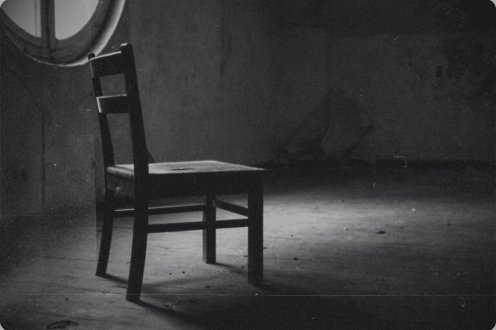 A single wooden chair sits in a worn, dimly lit room, bathed in soft light from a round window—quietly marking the absence of what once was.