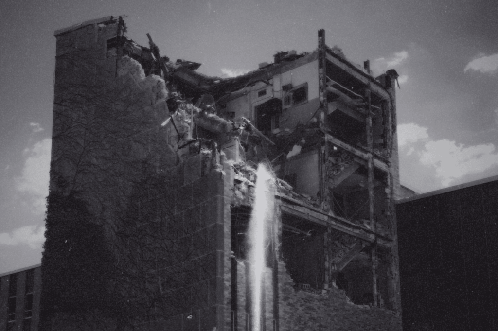 A black-and-white image of a partially collapsed building, its broken walls and spraying water evoking the raw aftermath of destruction and the haunting imprint of what once stood.