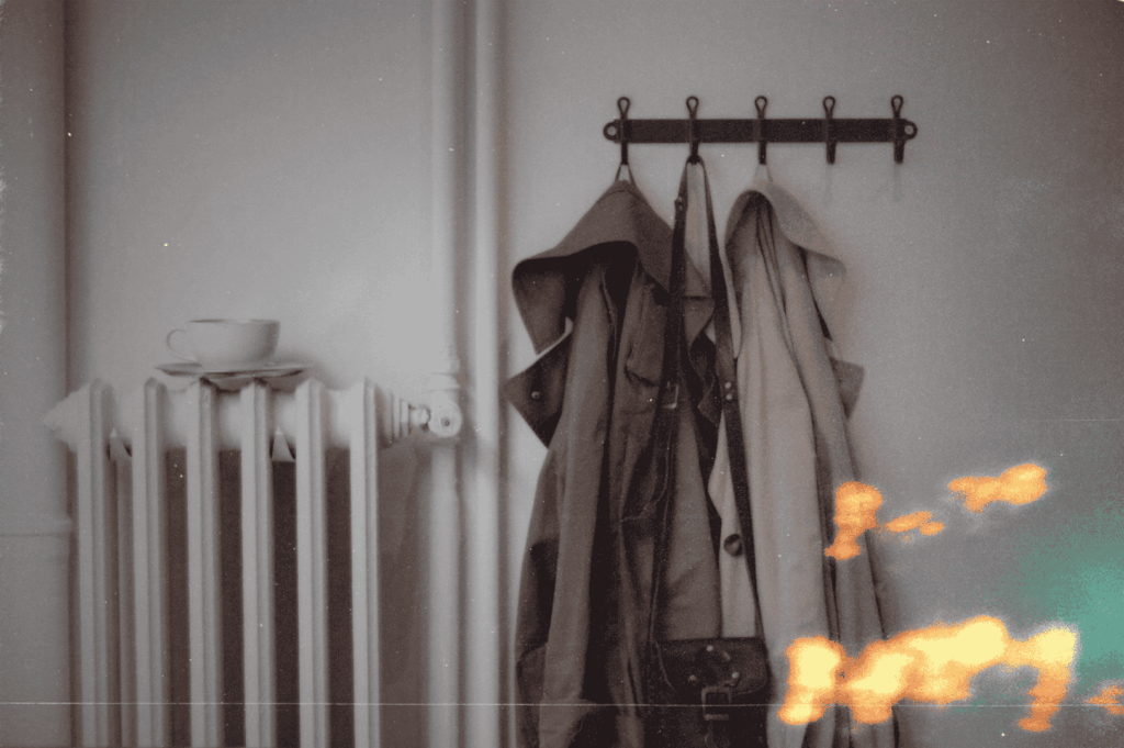 A row of muted coats hangs above a white radiator, where a lone teacup rests—quiet traces of daily life lingering in a quiet domestic space.