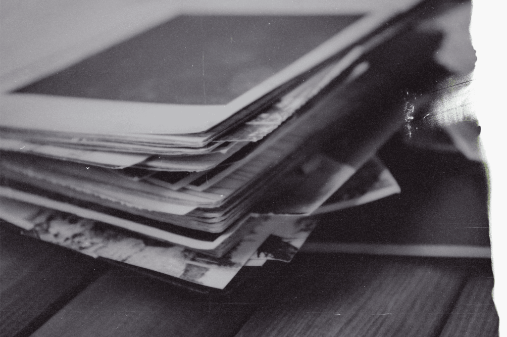 A black-and-white photograph of a disheveled stack of papers and notebooks on a wooden surface—an intimate archive of what once mattered, now quietly fading.