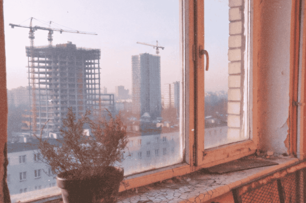A cracked windowsill with a lone potted plant overlooks rising towers—juxtaposing lived-in stillness with relentless urban renewal.