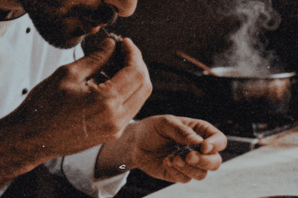 Close-up of a chef in white smelling fresh herbs at a stove, steam rising from a pot, suggesting careful tasting and memory-rich cooking