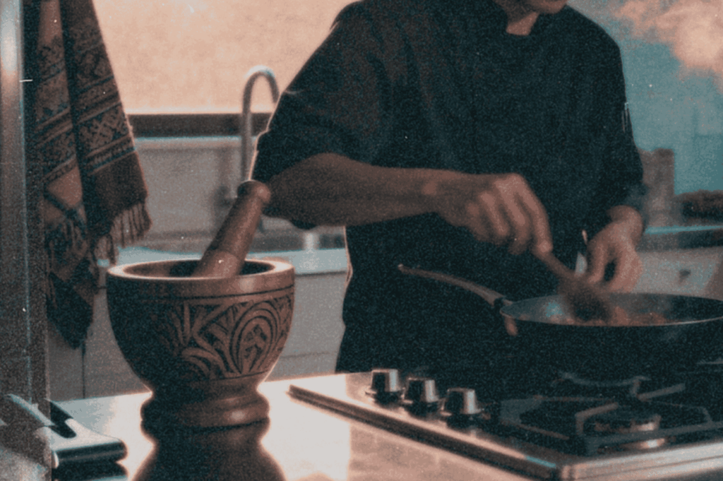A cook in a home kitchen stirring a steaming pan beside a carved mortar, reflecting traditional cooking methods.