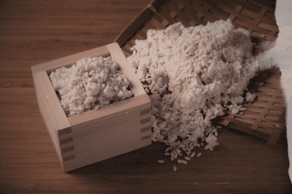 A traditional wooden masu box brimming with pearly koji rice sits beside a bamboo tray, grains spilling gently across a warm wooden surface—evoking the tactile rituals and ancestral care of Japanese fermentation.
