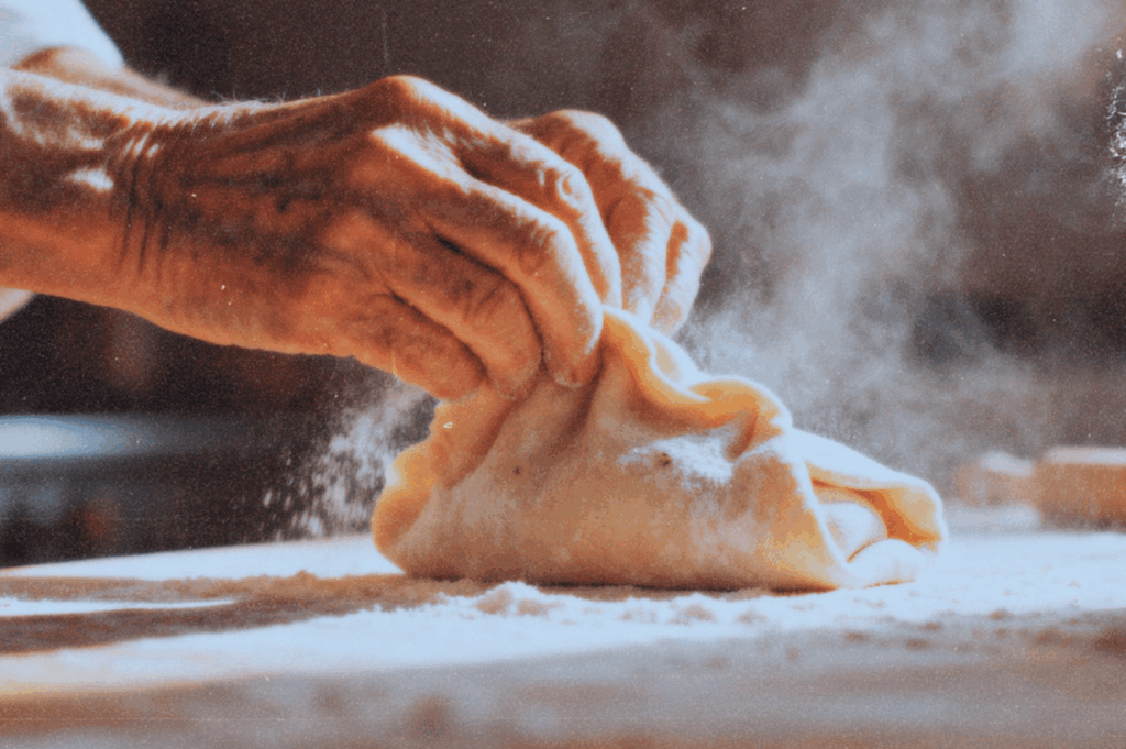Close-up of elderly hands folding dough on a floured table, flour dust rising in warm light.