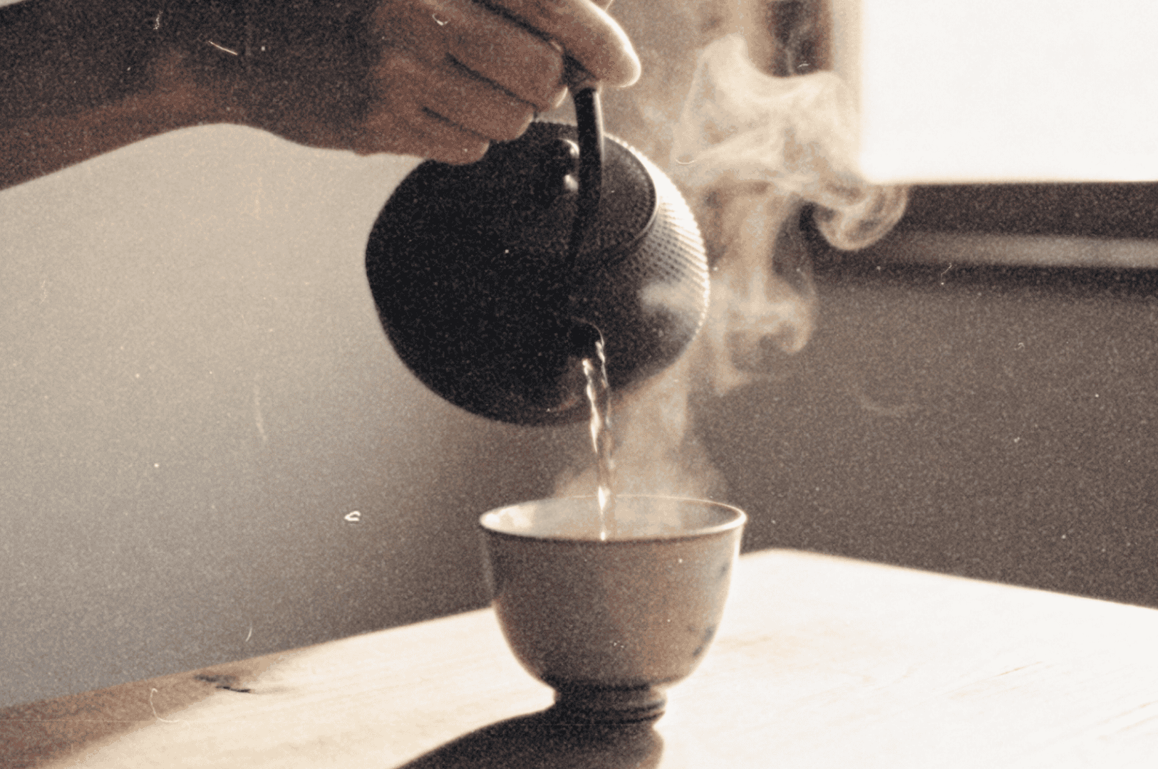 A hand pours hot tea from a small black teapot into a steaming cup on a sunlit wooden table.