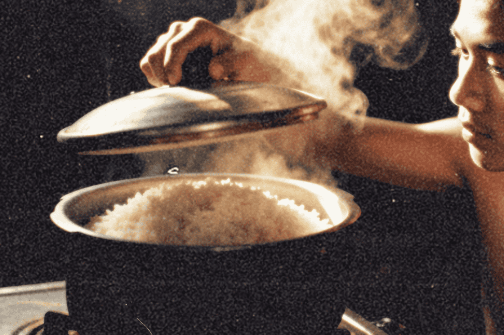 A person lifts the lid of a steaming pot filled with freshly cooked rice on a stove, with warm light highlighting the rising steam.
