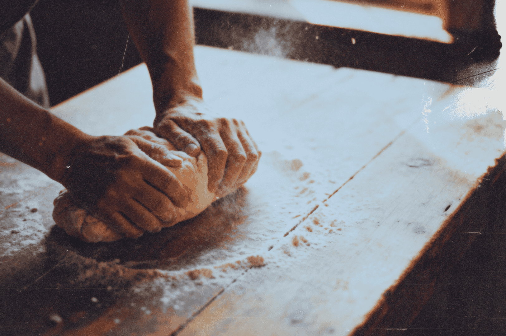 Hands kneading bread dough on a floured wooden table in warm light.