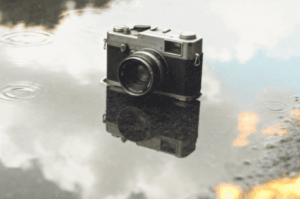 A vintage camera sits partially submerged in a puddle of water, reflecting the sky and clouds above. Raindrops ripple on the surface, creating a calm and artistic mood.