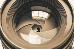  A close-up black and white image of a camera lens showing the aperture blades and a faint reflection of a person and a window in the glass.