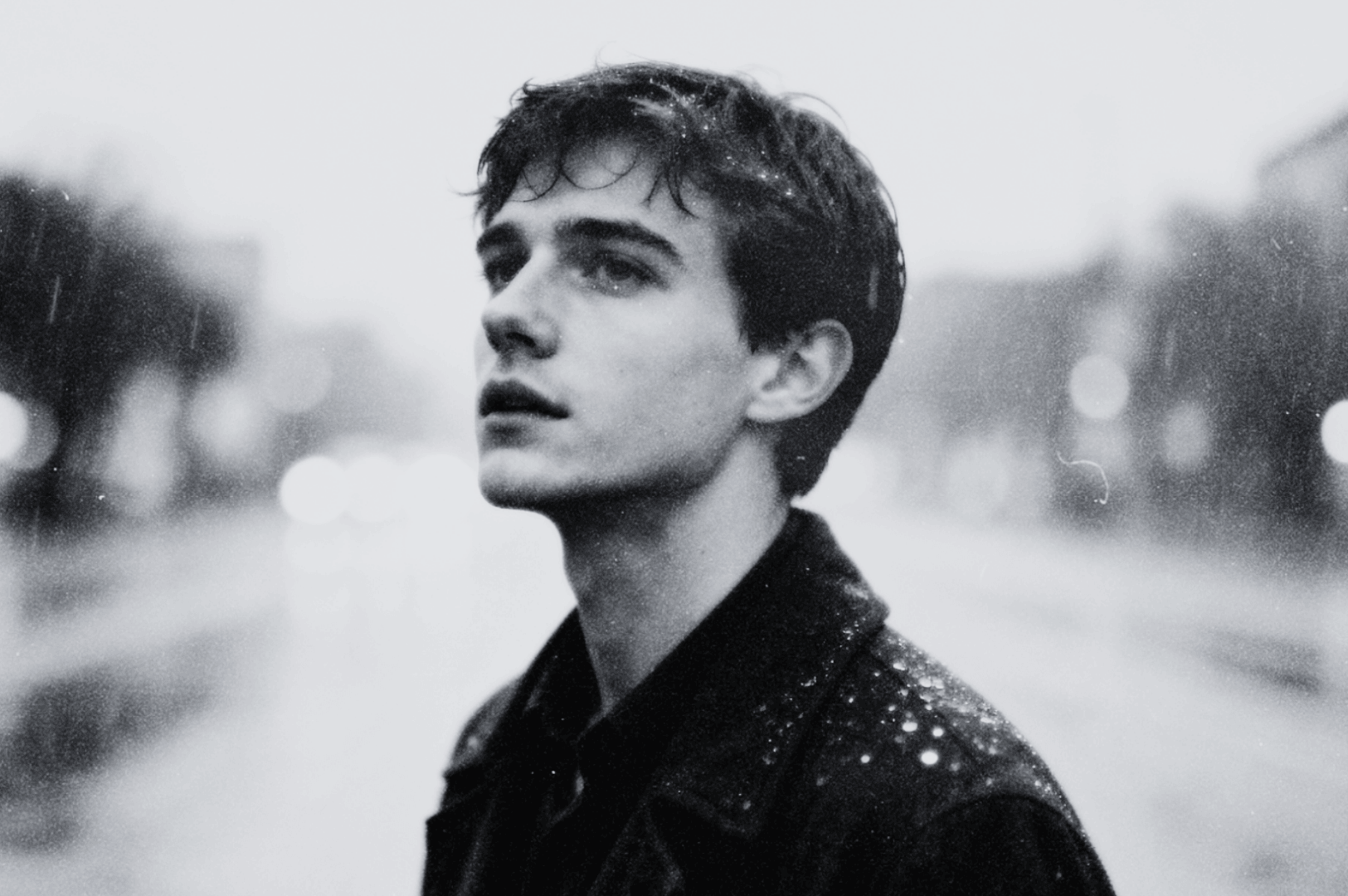 Black and white photo of a person standing in light rain on a blurry city street, looking upward.