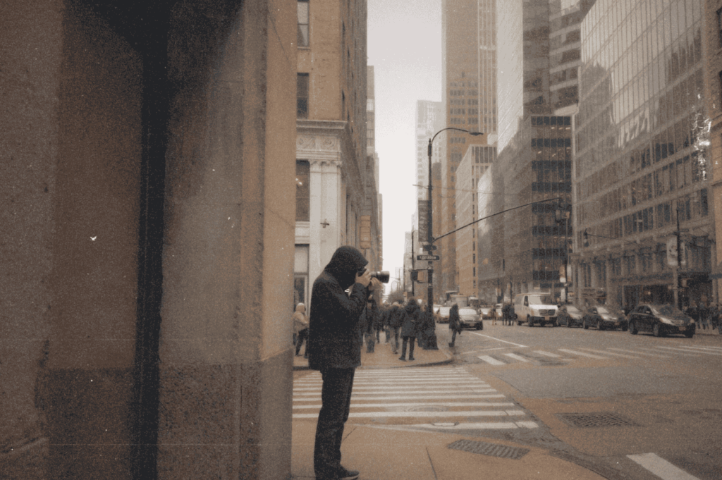 A photographer pauses at a busy city corner, framed by towering buildings and passing crowds—capturing a quiet moment of focus amid the metropolitan rush.