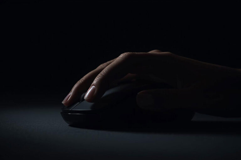 Close-up shot of a person's hand with manicured nails resting on a black computer mouse in a dark, low-key lighting setting, suggesting late-night work or focus.