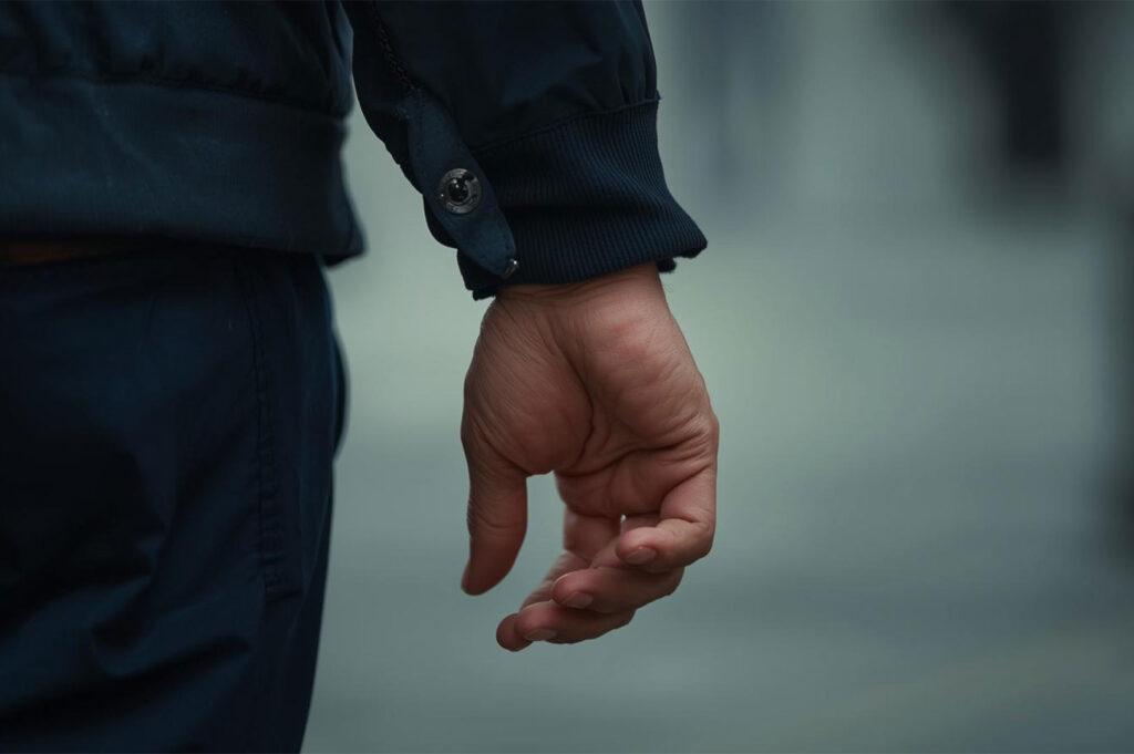 Close-up, cropped image of a man's right hand hanging down next to his body, wearing a navy blue jacket with a ribbed cuff and a button detail.