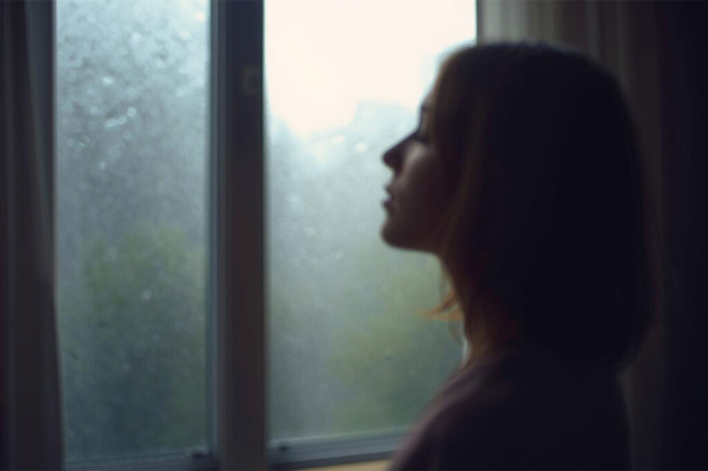 Silhouette of a young woman's profile looking out of a blurry window covered in raindrops, capturing a melancholic or introspective mood.