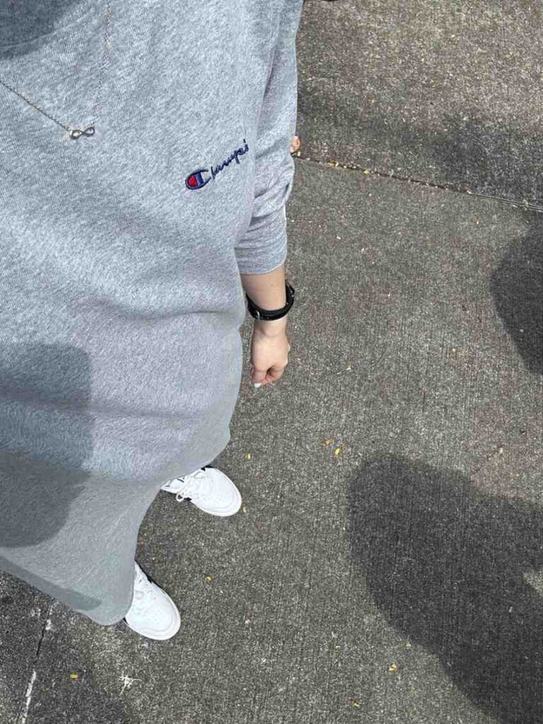 A high-angle shot shows a person standing on pavement, dressed in a grey Champion sweatshirt dress and white sneakers. Simple accessories, such as a silver infinity necklace and a black wristband, complement the casual attire.