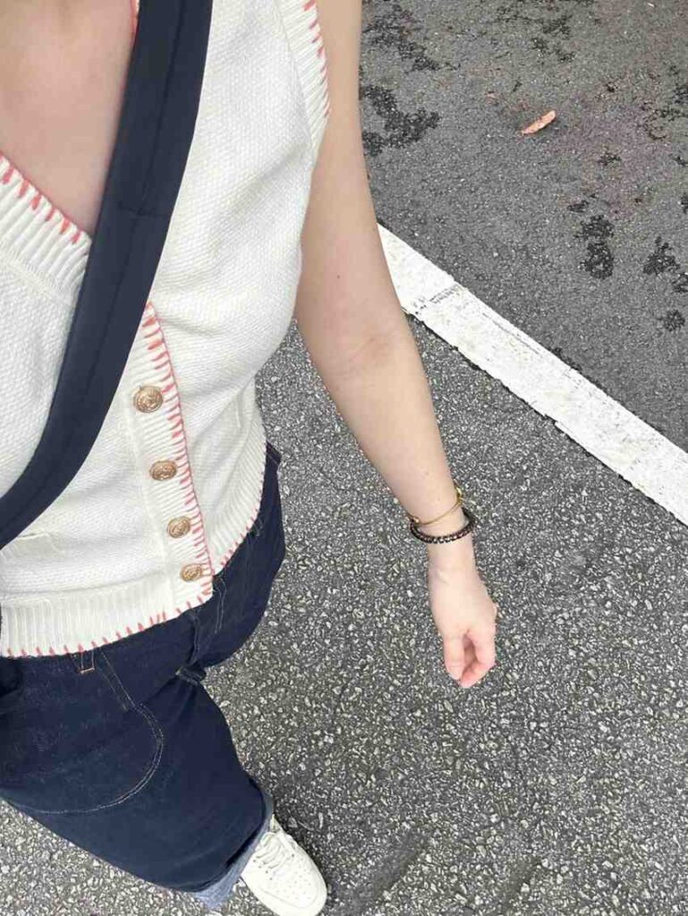 A person wears a sleeveless cream knit vest with coral stitching and gold buttons, paired with dark cuffed jeans and white sneakers. The casual outfit is accessorized with a black cross-body bag strap and bracelets, captured from a high-angle perspective against an asphalt surface.