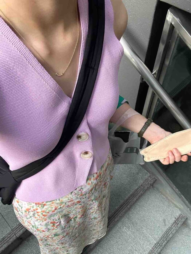 A person stands near a staircase railing, dressed in a sleeveless lavender cardigan top paired with a light-colored floral skirt. The outfit is accessorized with a simple gold necklace, a dark watch, and a black cross-body bag strap, while the individual holds a beige object in their hand.