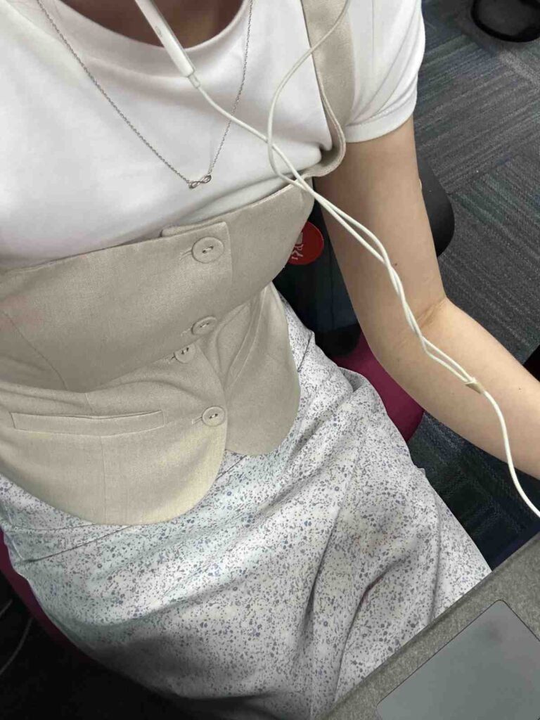 A person is seated wearing a layered outfit that combines a beige buttoned corset vest over a white t-shirt, paired with a light-colored floral skirt. White wired earphones and a delicate silver necklace with an infinity pendant complete the look, suggesting a relaxed yet stylish setting.