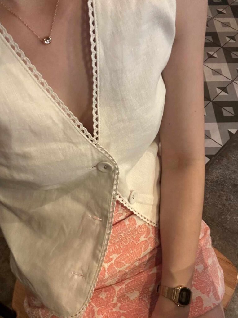 A person wears a cream-colored, sleeveless vest featuring scalloped lace trim, paired with a pink and white patterned skirt. The look is completed with gold accessories, including a delicate necklace and a digital wristwatch, captured from a high angle against a geometric tiled floor.