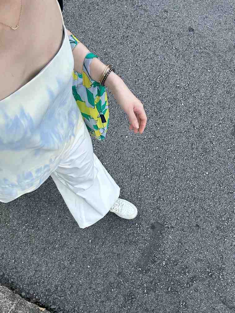 A high-angle shot captures a person wearing a strapless top with a blue tie-dye pattern paired with white wide-leg pants and sneakers. A vibrant tote bag featuring a lemon and leaf print hangs from their shoulder, complemented by simple gold jewelry and bracelets on their wrist.