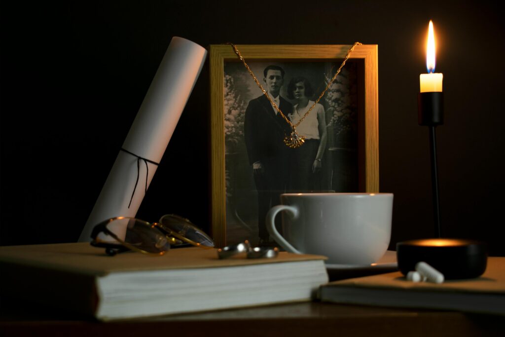 This moody still life features a framed black-and-white portrait of a couple, draped with a gold necklace and illuminated by the solitary flame of a candle.