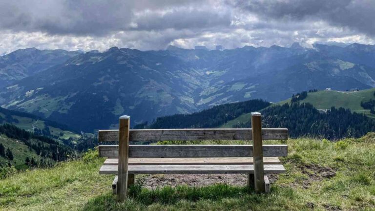 A wide-angle shot of a rustic wooden bench facing a vast, panoramic view of rolling blue and green mountain ranges under a dramatic, cloudy sky. The bench is positioned in the foreground on a grassy hill, overlooking a deep valley.