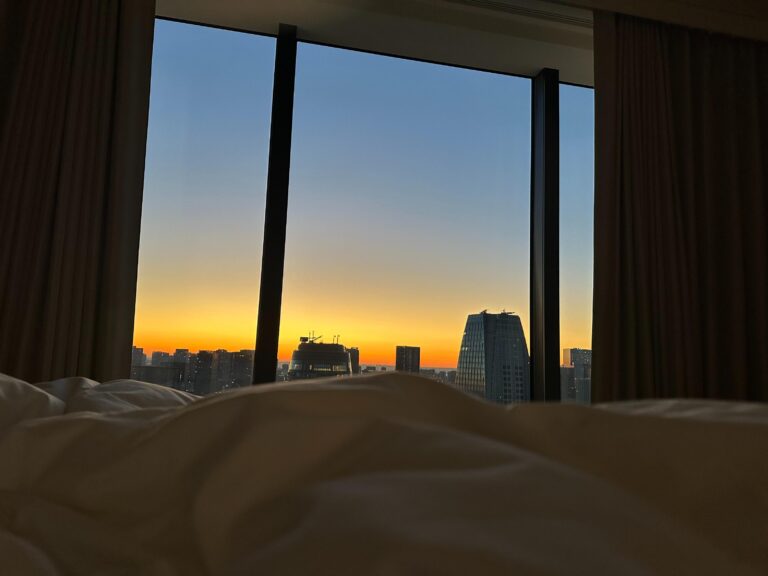 From the perspective of a bed with white linens, a large floor-to-ceiling window frames a silhouette of skyscrapers against a vibrant sky. The horizon glows with a gradient of deep orange and yellow fading into blue, suggesting a peaceful sunrise or sunset over the city.
