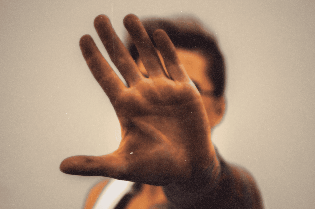 A sharply focused open hand blocks the camera, fingers splayed in quiet defiance, while the blurred figure behind it fades into anonymity—evoking the palm as both shield and storyteller.