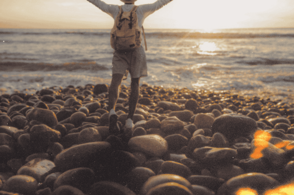 Silhouetted figure with arms raised on a rocky shore at sunset—embodying personal triumph, solitude, and the healing power of ocean horizons.
