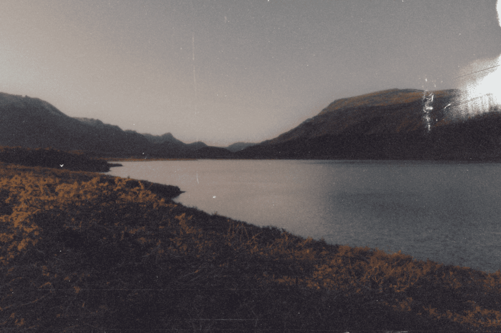  Vintage-toned lake bordered by softly lit mountains and a rugged grassy shore—evoking solitude, memory, and the quiet restoration found in nature’s embrace.