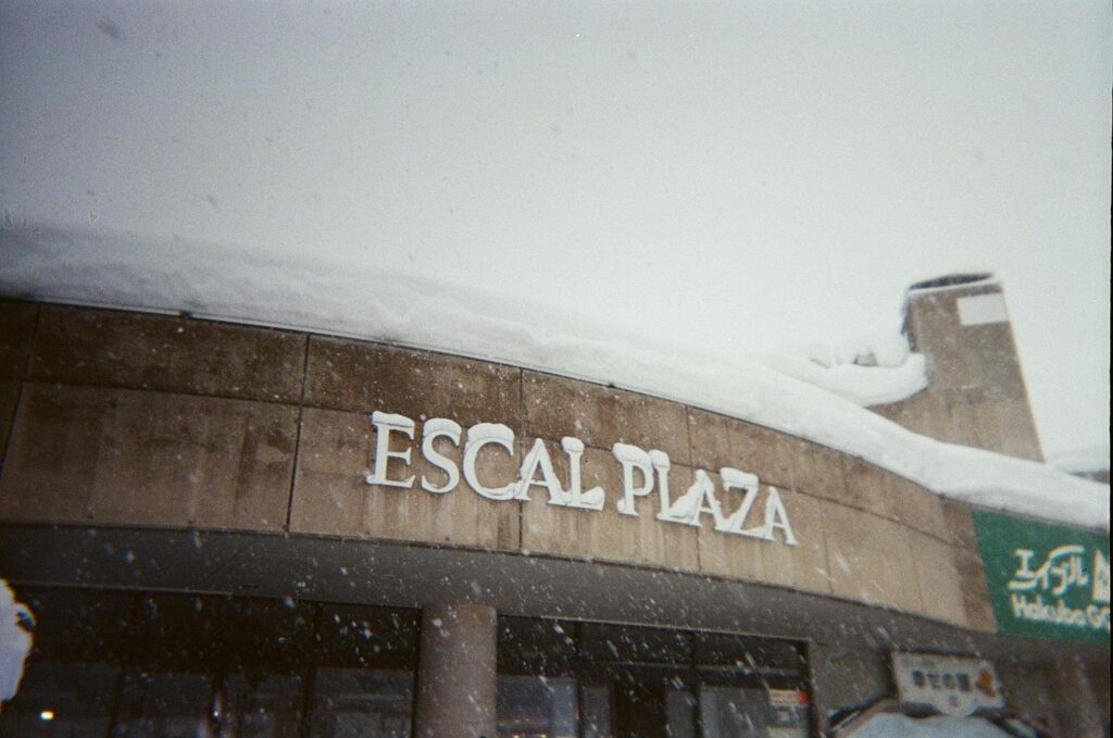 This grainy film photograph features the facade of a building labeled "ESCAL PLAZA," which is topped with a heavy, thick blanket of snow. Falling snowflakes obscure the grey sky, creating a moody and atmospheric winter scene typical of a ski resort environment.