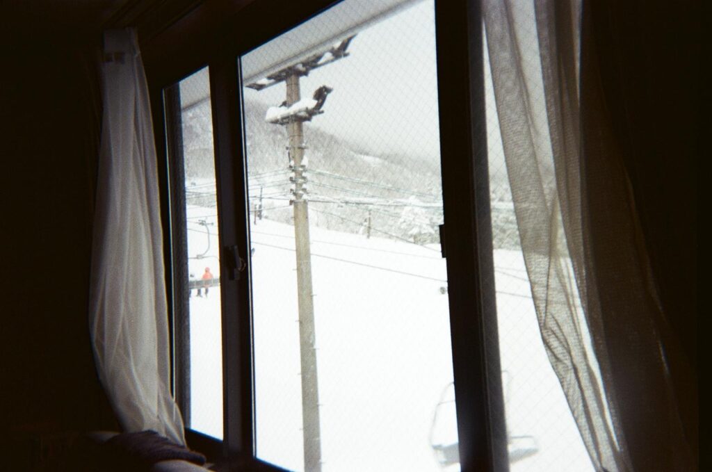 This film photograph frames a wintry view through a window, where dark frames and sheer curtains flank a look out onto a snowy ski slope. Outside, utility poles and chairlift cables cut across the white landscape, offering a cozy, interior perspective of the cold resort environment.