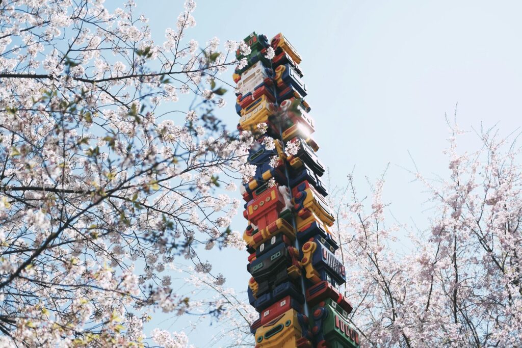 A towering, colorful sculpture constructed from stacked, robot-like forms stands framed by delicate cherry blossom branches.