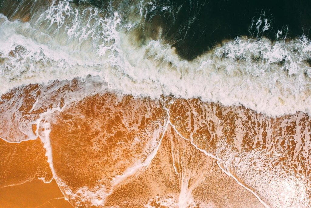 An aerial view captures the dynamic contrast between deep, teal-colored ocean waves and the vibrant golden-orange sand of the shoreline.