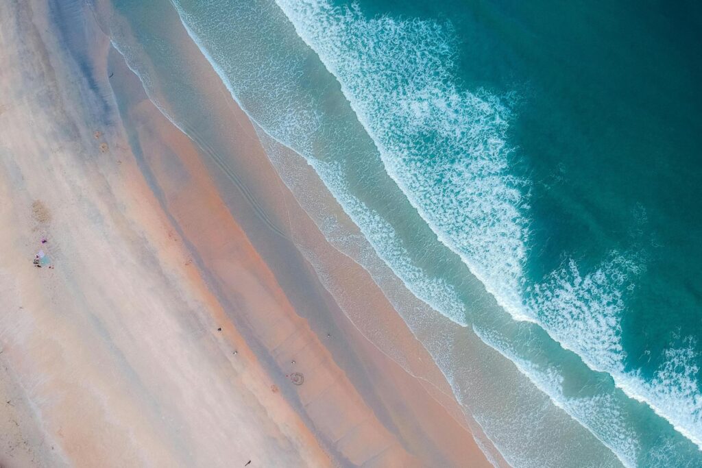 A high-angle aerial view depicts a vibrant diagonal coastline where layers of deep turquoise waves roll onto a smooth, sandy beach.