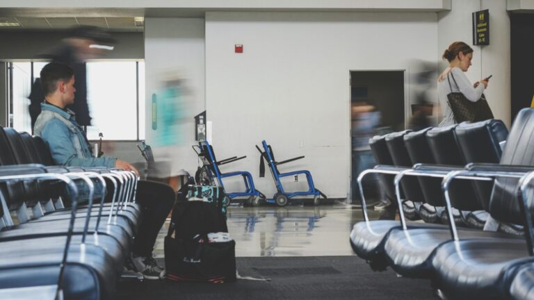 In a busy terminal, a man sits on a row of black chairs while a woman checks her phone, both appearing static amidst the blurred motion of passing people.