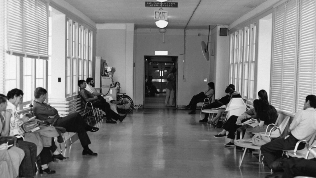 This black-and-white photograph depicts a long, sunlit corridor lined with people seated in chairs, suggesting a waiting area in a medical or institutional setting.