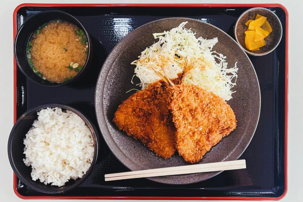 A Japanese set meal (teishoku) on a black and red tray. It features two pieces of golden-brown fried cutlet (likely tonkatsu or similar) served on a dark plate with a pile of shredded cabbage, a bowl of white rice, a bowl of miso soup, and a small dish of yellow pickled daikon radish (takuan)