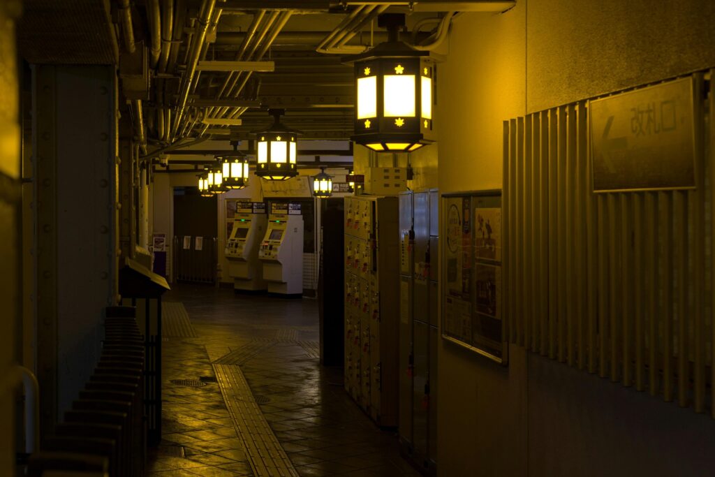 Warm, yellow light from decorative hanging lanterns illuminates a moody, industrial-style corridor, likely within a Japanese train station.