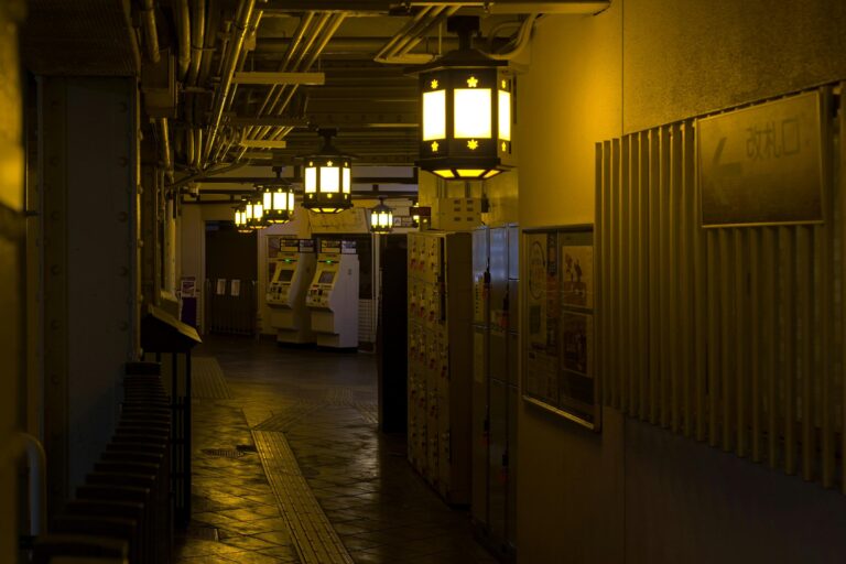 Warm, yellow light from decorative hanging lanterns illuminates a moody, industrial-style corridor, likely within a Japanese train station.