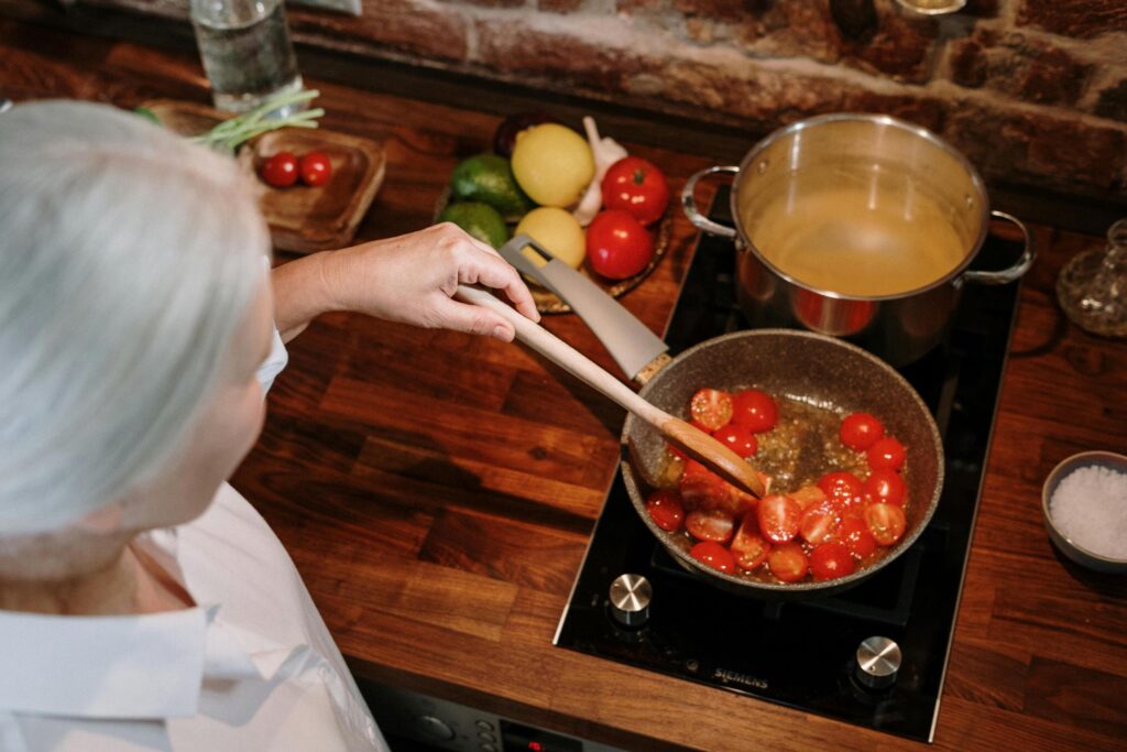 From a high angle, a woman with white hair is seen stirring cherry tomatoes in a frying pan on a black electric stovetop using a wooden spoon.