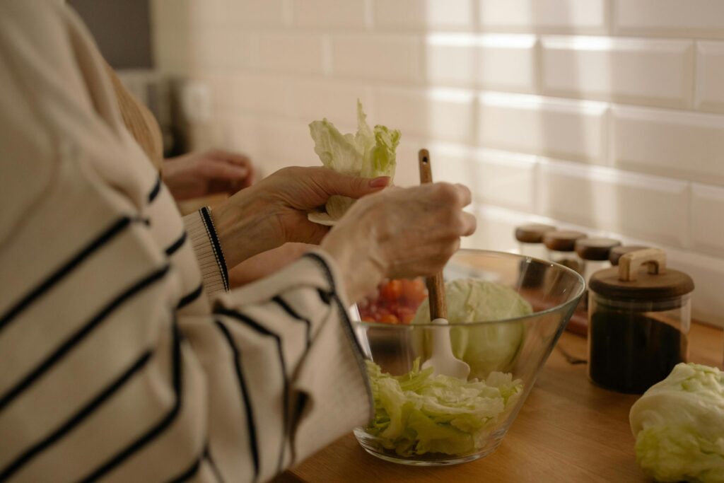 A person wearing a cream-colored striped sweater stands at a wooden counter, tearing fresh lettuce leaves by hand into a clear glass bowl.