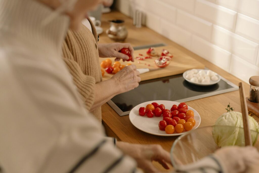 Two people prepare food at a wooden kitchen counter, where one person in a beige sweater arranges red and orange cherry tomatoes on a white plate.