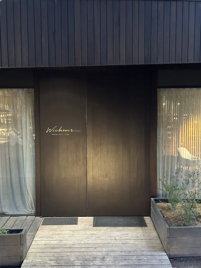 This image depicts the stylish entrance to "Wickens at the Royal Mail Hotel," defined by a dark, vertical-paneled exterior and gold signage on the central door. Flanked by curtained windows, a simple wooden boardwalk leads the way to the entrance, creating a refined and welcoming atmosphere.