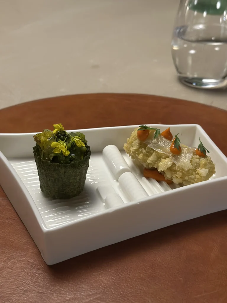 This image features a duo of intricate appetizers served on a ridged white rectangular dish. On the left is a dark green tartlet filled with vegetables and yellow flowers, while the right displays a crispy fried cylinder garnished with orange puree and herbs.