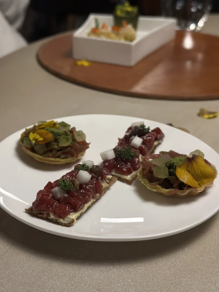 This image showcases a selection of delicate canapés, including rectangular crisps topped with diced red tartare and white cubes, alongside round tartlets adorned with yellow edible flowers. The appetizers are meticulously arranged on a white plate, set against a blurred background that implies a sophisticated multi-course dining experience.