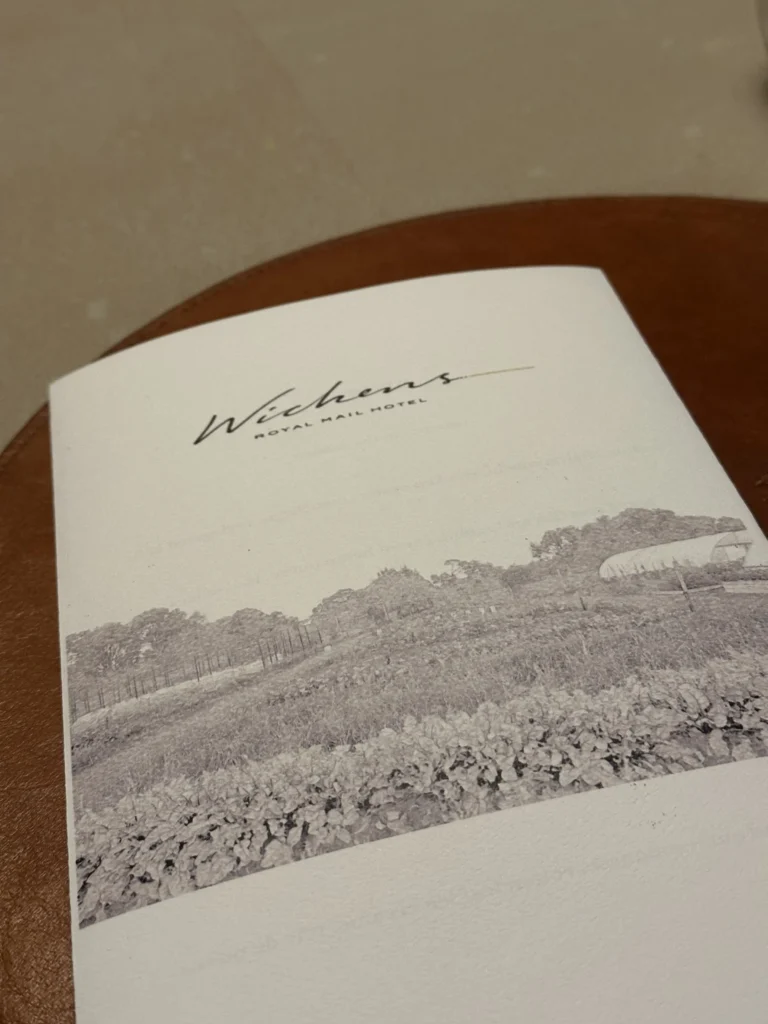 This image displays the cover of a menu for "Wickens at the Royal Mail Hotel," characterized by elegant script typography above a monochrome photograph of a garden. The visual depicts rows of leafy crops and a greenhouse structure, emphasizing a connection to fresh produce and the outdoors.